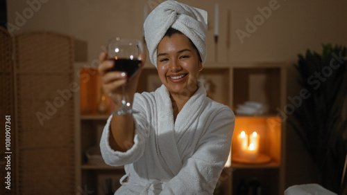 Obraz na plátně A beautiful young hispanic woman in a spa wearing a white robe and towel on her head, holding a glass of red wine and smiling in a dimly lit room with candles in the background