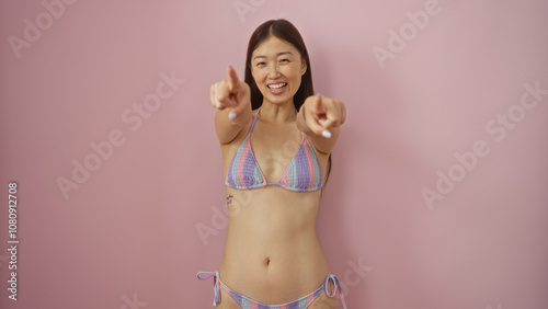 Young, beautiful, asian woman in a colorful bikini points and smiles cheerfully at the camera against an isolated pink background