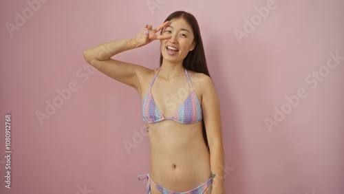 Woman posing in a colorful bikini against an isolated pink background, showing a peace sign and smiling, young, beautiful, chinese, and attractive adult