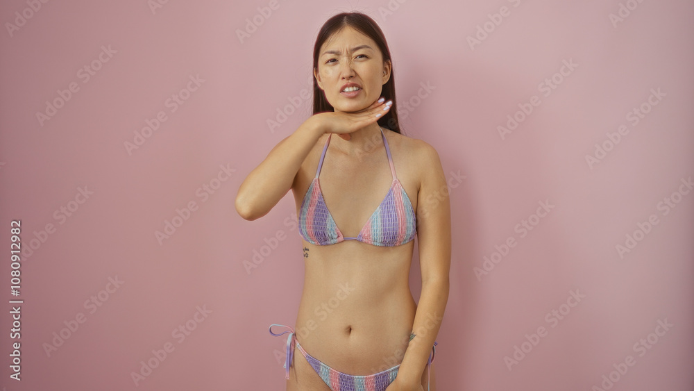 Beautiful young asian woman in a bikini makes a neck-cutting gesture against an isolated pink background, conveying frustration or discontent. Beautiful young asian woman in a bikini makes a neck-cutting gesture against an isolated pink background, conveying frustration or discontent.