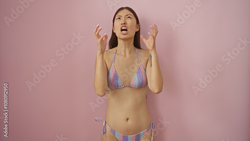 Attractive young asian woman expressing frustration in a bikini on an isolated pink background