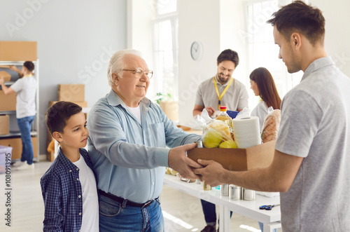 Friendly male volunteer holding box with essentials, giving free food donation to poor refugee family, smiling grandfather and grandson in charitable foundation. Humanitarian help and social support. photo