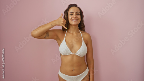 Beautiful young hispanic woman making call-me gesture while standing in bikini against isolated pink background