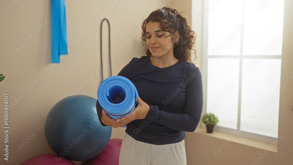 Young, attractive, hispanic woman holding a yoga mat at a gymnasium indoor sport center, with gym equipment and natural lighting. Young, attractive, hispanic woman holding a yoga mat at a gymnasium indoor sport center, with gym equipment and natural lighting.