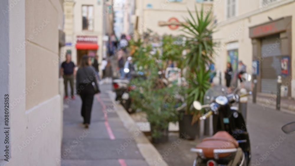Obraz premium Blurred street in marseilles with woman walking along urban sidewalk surrounded by defocused buildings and plants in soft sunlight, creating a bokeh effect.