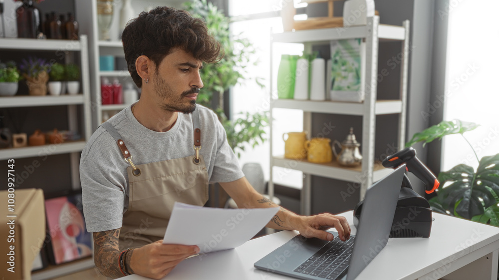 Young man with a beard holding a document while using a laptop in a well-decorated shop with shelves full of various home decor items. Young man with a beard holding a document while using a laptop in a well-decorated shop with shelves full of various home decor items.