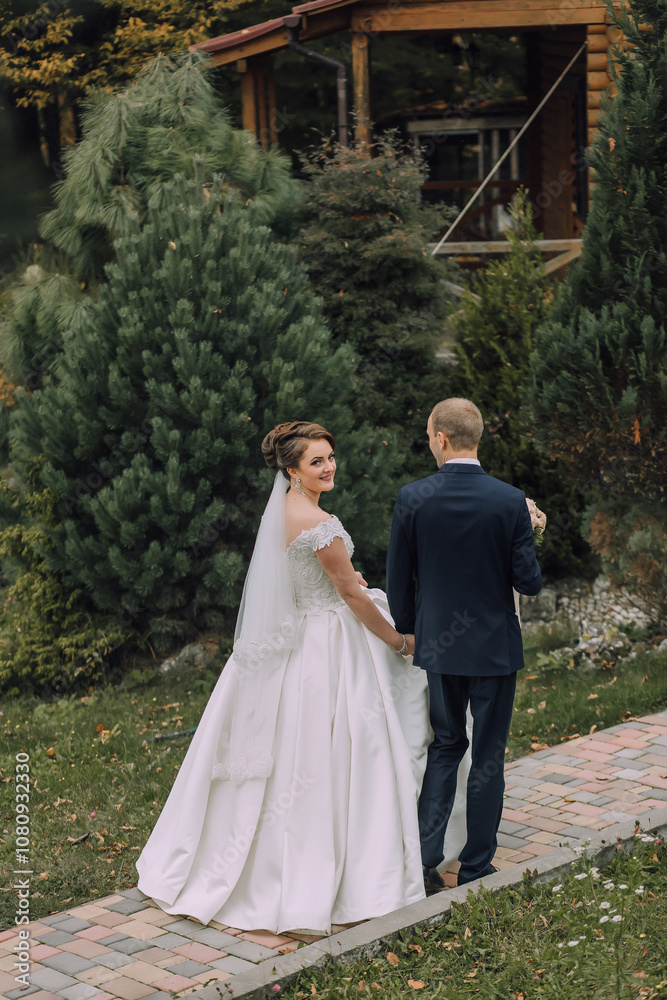 A bride and groom are walking down a brick path in front of a house. The bride is wearing a white dress and a veil, while the groom is wearing a blue suit. They are holding hands and smiling A bride and groom are walking down a brick path in front of a house. The bride is wearing a white dress and a veil, while the groom is wearing a blue suit. They are holding hands and smiling