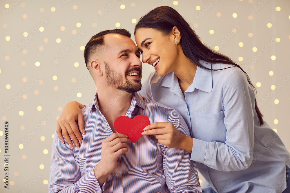 Portrait of a young happy smiling man and woman hugging and holding red paper heart in hands standing indoors on lights bokeh background. Happy couple in love. Valentines day concept. Portrait of a young happy smiling man and woman hugging and holding red paper heart in hands standing indoors on lights bokeh background. Happy couple in love. Valentines day concept.