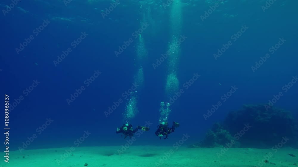 Pair of scuba divers swim over sandy bottom, approaching and waving greetings, in the blue depths sea, Slow motion of Scuba divers swim in the deep, a long trail of air bubbles rising above them