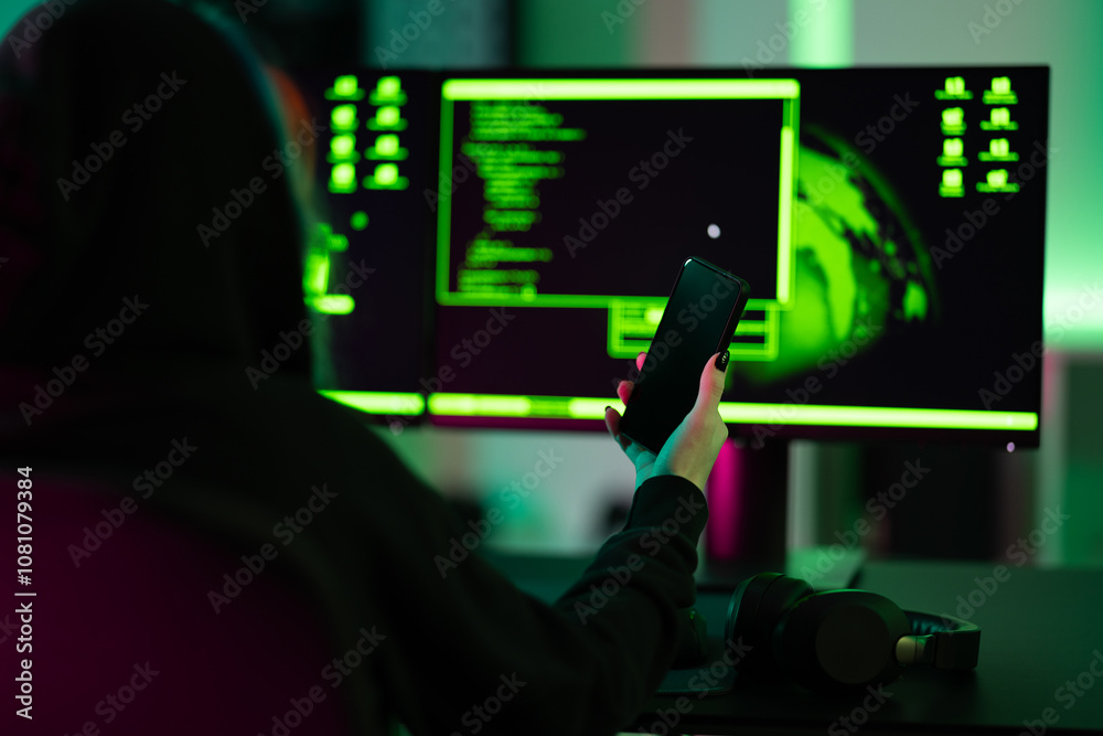 Hacker holding smartphone in front of dual screens with coding ...