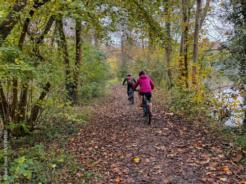 Two cyclists riding through a scenic forest trail covered in autumn leaves near a tranquil river