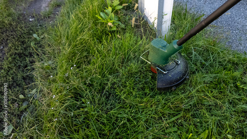 A green grass trimmer cuts overgrown lawn edges, symbolizing summer garden maintenance and landscaping challenges
