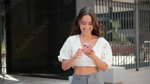 Slow motion. Hispanic young woman walking outdoors using a cellphone, smiling while texting or browsing on her smartphone. Latin teenage girl using a smart phone to send messages at the street
