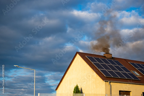 Black smoke from the chimney of a single-family home. Roof covered with solar panels. Roof against a blue, slightly cloudy sky.