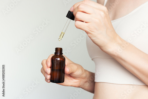 Profile shoot of young woman in beige underwear holding in her hand in front of her dark glass bottle of cosmetic liquid and dripping into it with pipette. Skin care. Isolated on a beige background