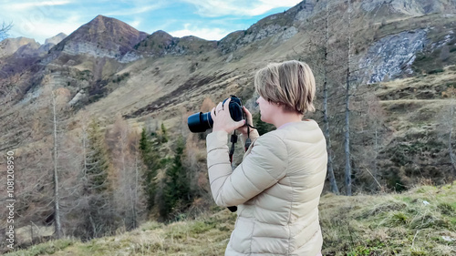 A woman in a beige jacket captures scenic mountain views with her camera, ideal for adventure travel and nature photography concepts