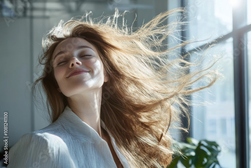 A joyful woman with a broad smile stands by a window with her hair flowing freely in the breeze, illuminated by soft natural light, embodying pure happiness. photo