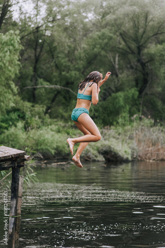 Athletic beautiful girl, adult child jumping from a wooden bridge, pier into the water on a lake outdoors in nature in summer. Photography, portrait, lifestyle, outdoor concept, sport.