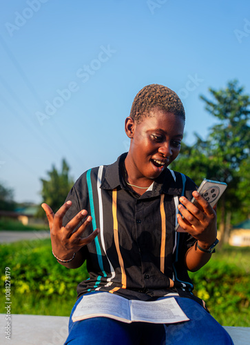 Black African female student sitting with a book and a mobile phone smiling open hands in excitement using phone winning happy looking at her phone in excitement afro low haircut chocolate skin