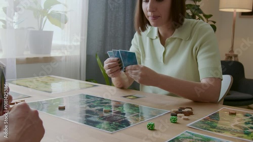 Over the shoulder view of young woman rolling dice, showing card to friend and making move while playing tabletop game during day at home photo