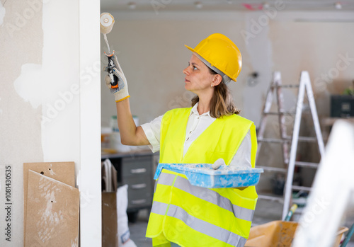 Professional female painter in protective helmet and yellow vest paints the walls with a paint roller photo