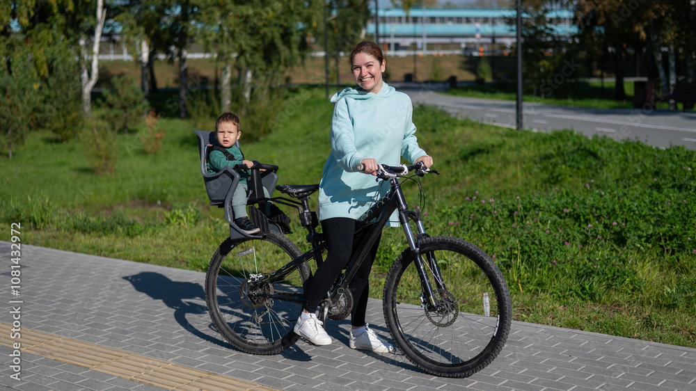 Fototapeta premium Caucasian woman riding bicycle with toddler son sitting behind her in child seat in park. 