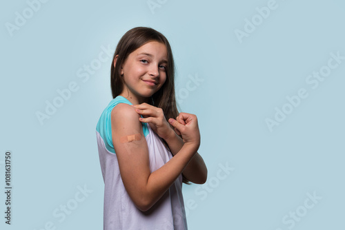 A young girl shows off her vaccinated arm by flexing and smiling, proudly wearing a bandage