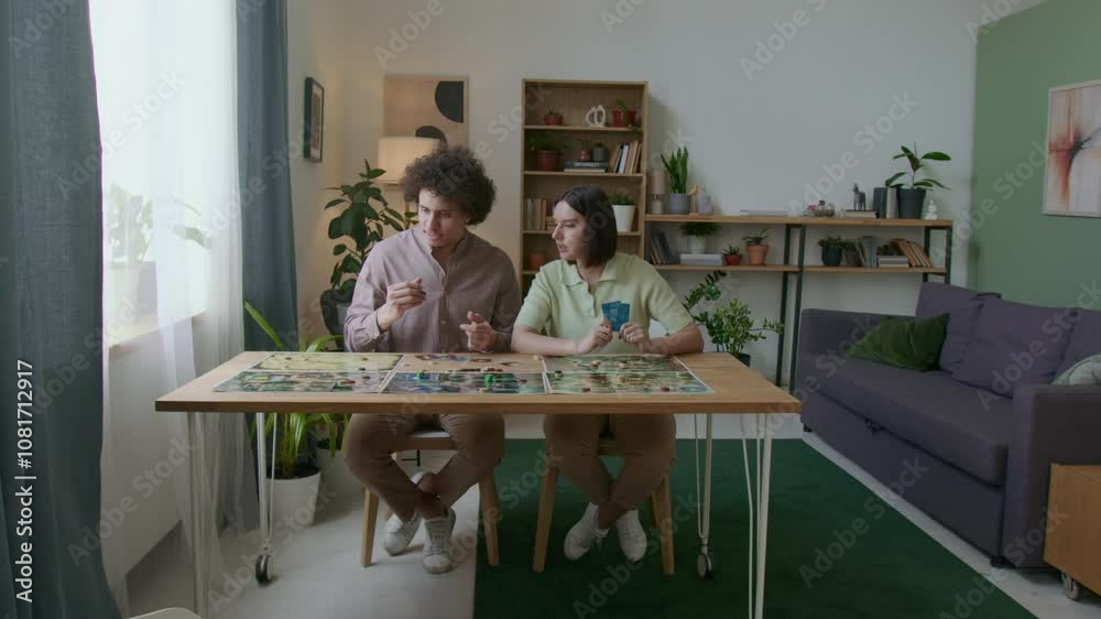 Young couple sitting at table with game boards and tokens spread out, discussing cards and making moves while playing tabletop game in cozy modern living room Young couple sitting at table with game boards and tokens spread out, discussing cards and making moves while playing tabletop game in cozy modern living room