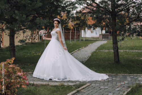 A woman in a white wedding dress is standing in front of a house. She is holding a bouquet of flowers and a veil. The scene is peaceful and serene, with the woman looking happy and content