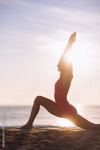 A silhouette of a woman in pink activewear performs a yoga stretch at sunset on the beach. The serene ocean backdrop and warm glow of the setting sun create a tranquil, mindful atmosphere, perfect for