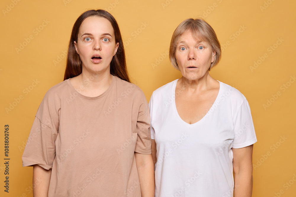 Two women with surprised expressions stand side by side against a bright yellow background in a studio setting, showcasing a moment of shared astonishment Two women with surprised expressions stand side by side against a bright yellow background in a studio setting, showcasing a moment of shared astonishment