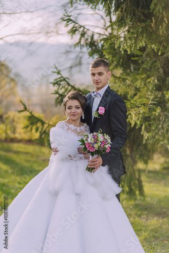 A bride and groom are posing for a picture in a forest. The bride is wearing a white dress and a fur stole, while the groom is wearing a suit. They are holding flowers and a bouquet photo