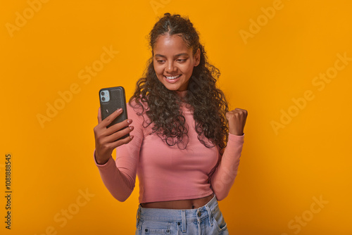 With a bright smile and a raised fist, a young Indian woman captures her excitement on a smartphone, set against a lively yellow background that radiates positivity and energy.