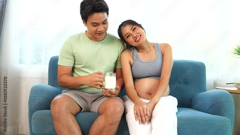 Asian husband takes care of his seven-month pregnant wife by giving her milk from a glass for healthy nutrition and calcium. Pregnant women sit together on the sofa in the living room at home. Asian husband takes care of his seven-month pregnant wife by giving her milk from a glass for healthy nutrition and calcium. Pregnant women sit together on the sofa in the living room at home.