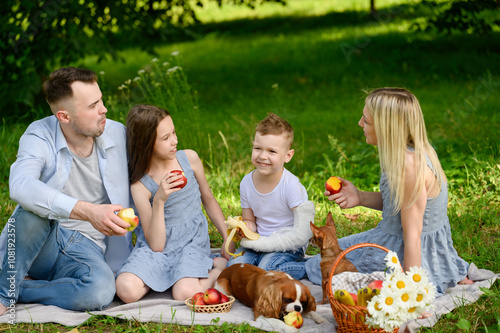 Family picnic in park. Happy parents with children enjoy communication, eat fruits during picnic on lawn, dog eat apple. Family outdoor recreation concept, family communication photo