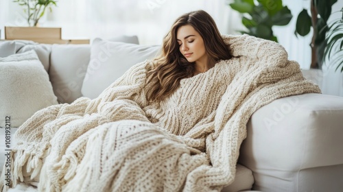 A young woman is relaxing on a white sofa, wrapped in a cozy blanket. She is looking at something off-camera, with a serene expression.