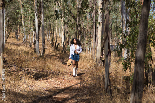 Latin woman, young, beautiful, dark-haired, with curly hair, with a white shirt and top, denim skirt and hat, walking alone and calmly through a eucalyptus forest. Concept peace, relaxation.