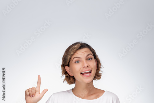 A young woman with short curly hair and a white shirt points upward with a joyful expression, isolated on a white background. Her enthusiastic smile and lively gesture make this image ideal for