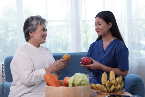 Asian nutritionist nurse holding fruit recommending healthy food to gray-haired senior woman chatting on sofa in hospital lounge photo