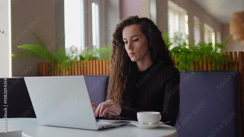Young woman with long, curly brown hair, wearing a black sweater, working on a laptop in a cozy, modern cafe. She is focused on her work, with a cup of coffee on the table. Green plants and soft light Young woman with long, curly brown hair, wearing a black sweater, working on a laptop in a cozy, modern cafe. She is focused on her work, with a cup of coffee on the table. Green plants and soft light