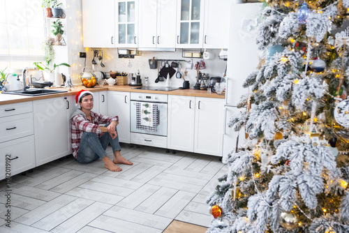 A lonely sad and tired man in a Santa hat is sitting on the floor of a festive white kitchen with Christmas decor photo