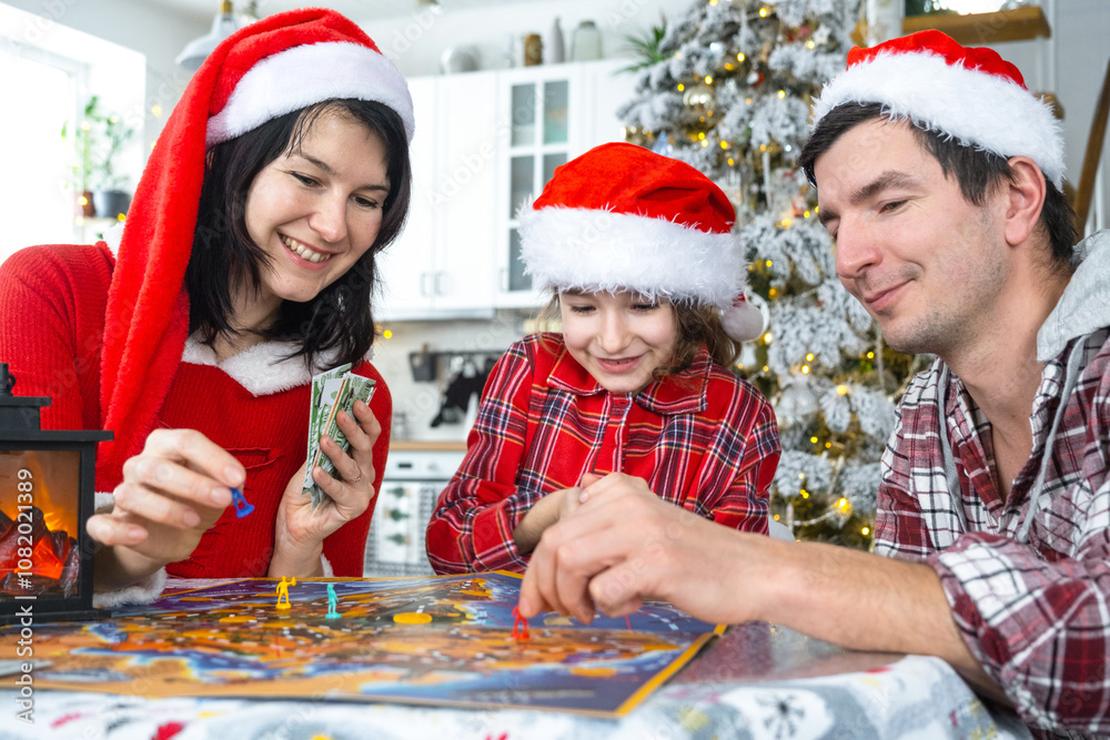 Happy traditional family of dad, mom, daughter in festive Santa hats Playing board game at table in a white kitchen with a Christmas tree and decor. New Year, family values. Happy traditional family of dad, mom, daughter in festive Santa hats Playing board game at table in a white kitchen with a Christmas tree and decor. New Year, family values.