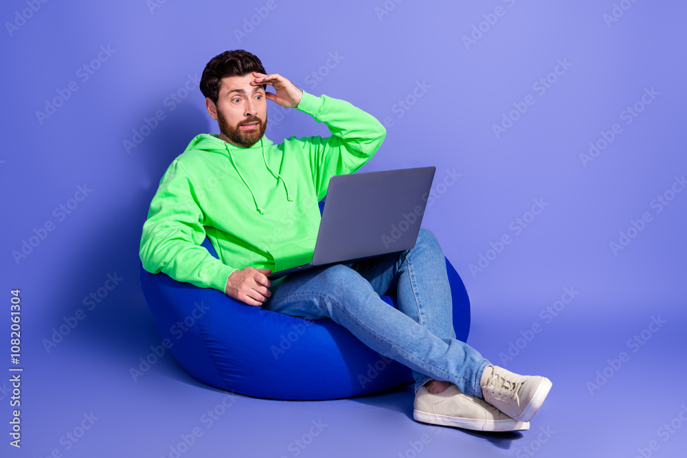 Full size photo of pretty young male sit beanbag hold netbook confused wear trendy green outfit isolated on purple color background