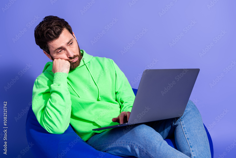 Photo portrait of pretty young male sit beanbag hold netbook boring tired wear trendy green outfit isolated on purple color background Photo portrait of pretty young male sit beanbag hold netbook boring tired wear trendy green outfit isolated on purple color background