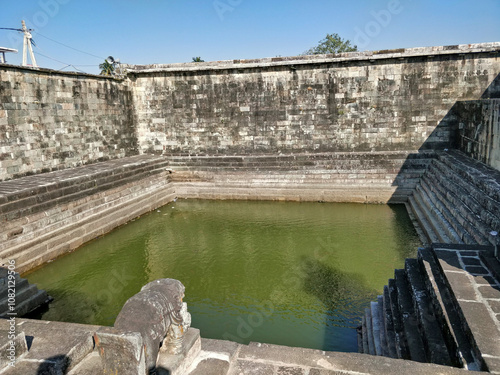 The temple tank at Chennakeshava Temple, Belur, features stone walls with intricate carvings, steps leading to green reflective water, and serene surroundings, showcasing Hoysala architectural beauty. photo