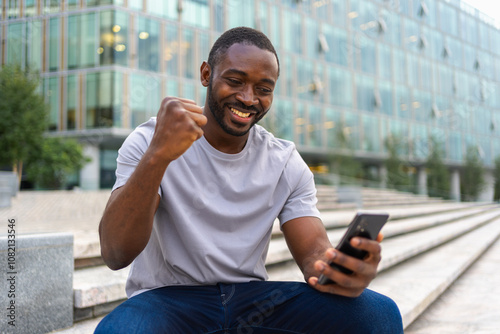 Fotografía Happy African American man euphoric winner with smartphone on street in city