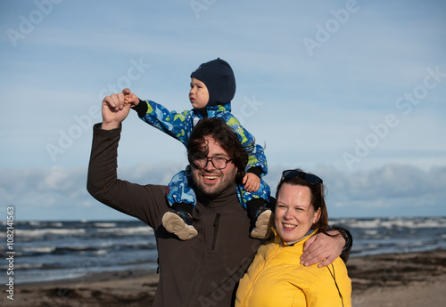 A happy family strolls along a chilly autumn beach, with their young son joyfully running ahead, creating lasting memories as the crisp wind and soothing waves embrace them in a moment of pure photo