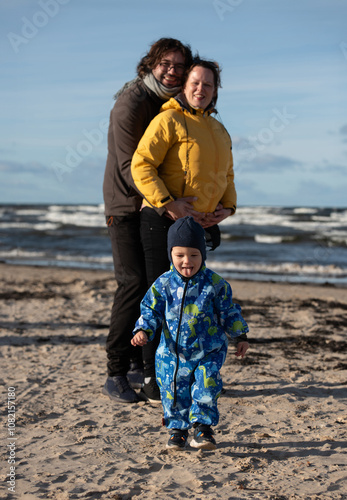 A happy family strolls along a chilly autumn beach, with their young son joyfully running ahead, creating lasting memories as the crisp wind and soothing waves embrace them in a moment of pure photo