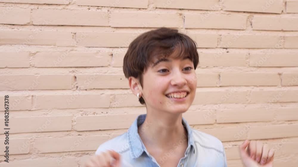 A Cheerful Smiling Young Individual with Short Hair Posing in Front of a Light Brick Wall A Cheerful Smiling Young Individual with Short Hair Posing in Front of a Light Brick Wall
