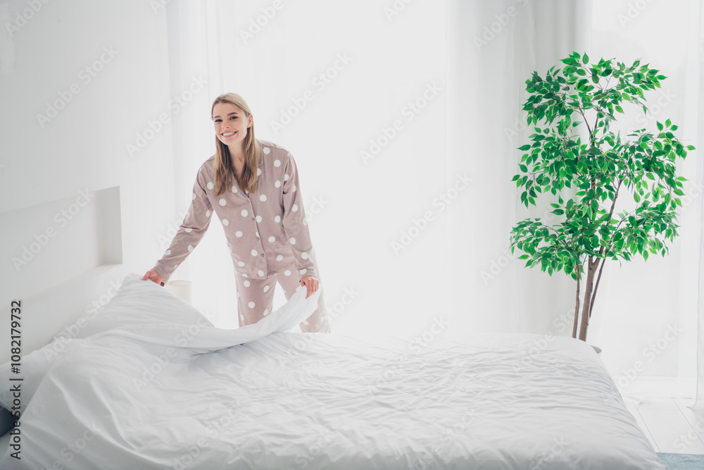 Fototapeta premium Young woman in cozy polka dot pajamas making a comfortable bed in airy bedroom with natural daylight
